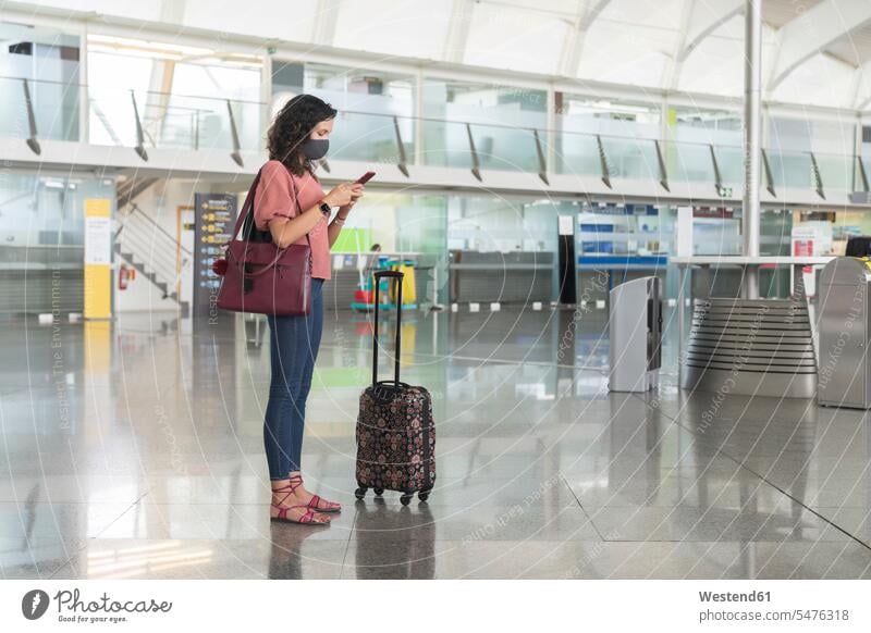 Junge Frau mit schützender Gesichtsmaske, die am Flughafen stehend Textnachrichten auf einem Smartphone verschickt Farbaufnahme Farbe Farbfoto Farbphoto