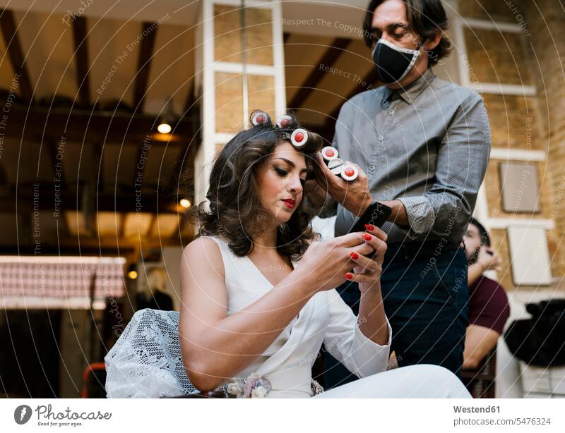Männlicher Friseur frisiert während die Braut während der Pandemie ein Smartphone im Salon benutzt Farbaufnahme Farbe Farbfoto Farbphoto Innenaufnahme