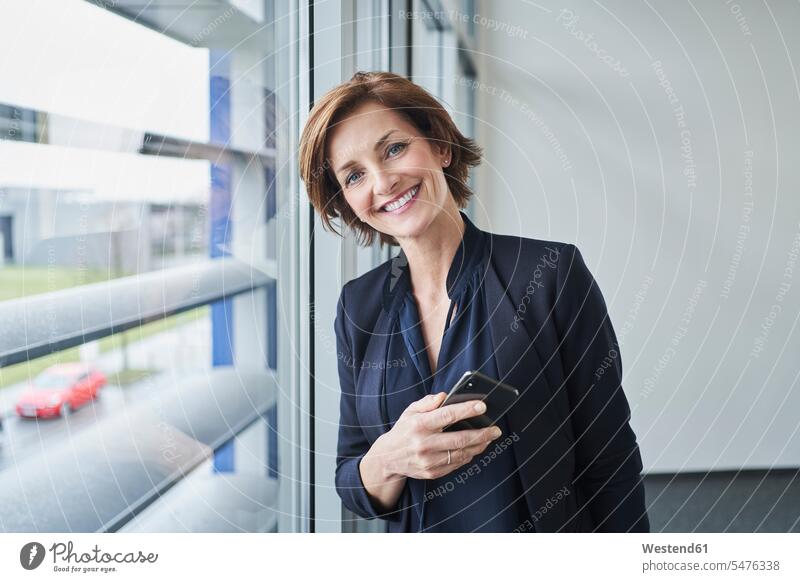Porträt einer selbstbewussten Geschäftsfrau am Fenster mit Handy in der Hand Job Berufe Berufstätigkeit Beschäftigung Jobs geschäftlich Geschäftsleben
