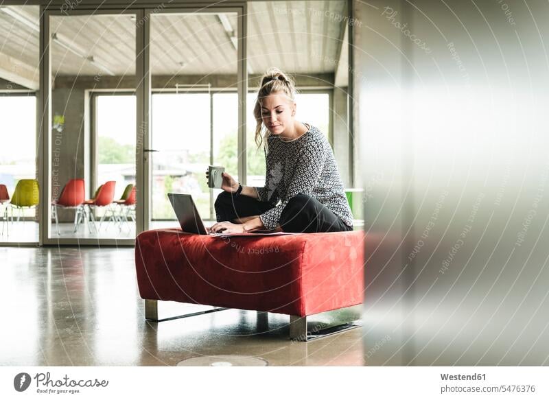 junge Geschäftsfrau, die in einem modernen Büro arbeitet, auf einem Hocker sitzt und einen Laptop benutzt Office Büros Schneidersitz Geschäftsfrauen