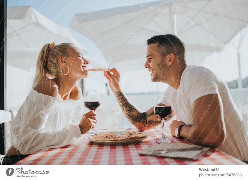 Glücklicher junger Mann gibt seiner Freundin Pizza zu essen, während er im Restaurant sitzt Farbaufnahme Farbe Farbfoto Farbphoto Spanien Freizeitbeschäftigung