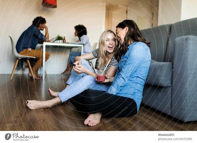 Zwei fröhliche junge Frauen sitzen auf dem Boden mit Handy und Getränk und Freunde im Hintergrund Freundinnen Böden Boeden weiblich Fröhlichkeit Frohsinn