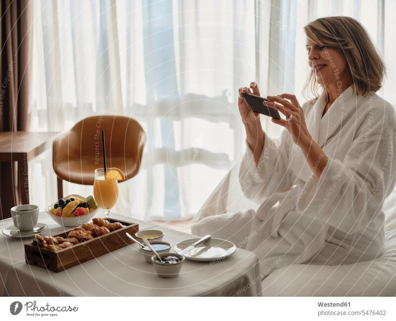 Lächelnde blonde ältere Frau fotografiert Frühstück über Smartphone, während sie im Hotelzimmer auf dem Bett sitzt Farbaufnahme Farbe Farbfoto Farbphoto