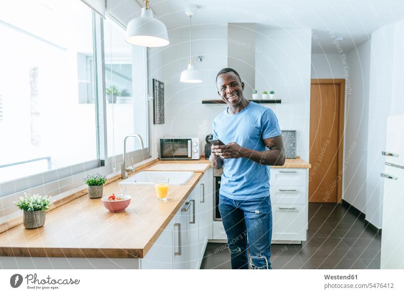 Porträt des glücklichen jungen Mannes mit Handy in der Küche zu Hause Leute Menschen People Person Personen Afrikanisch Afrikanische Abstammung dunkelhäutig