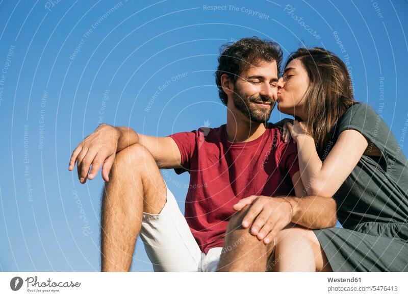 Junge Frau küsst Mann, während sie vor klarem Himmel sitzt Farbaufnahme Farbe Farbfoto Farbphoto Außenaufnahme außen draußen im Freien Tag Tageslichtaufnahme