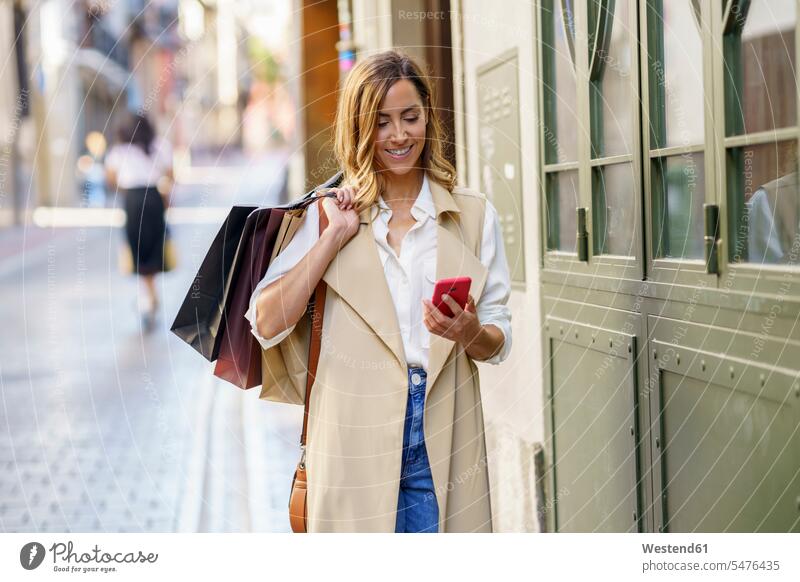 Frau mit Einkaufstasche, die ein Mobiltelefon benutzt, während sie am Fussweg in der Stadt steht Farbaufnahme Farbe Farbfoto Farbphoto Außenaufnahme außen
