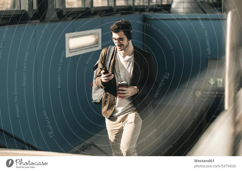 Lächelnder junger Mann mit Kopfhörern, Mobiltelefon und Kaffee zum Mitnehmen beim Spaziergang am Bahnhof lächeln Bahnhöfe Bahnhoefe Männer männlich gehen gehend