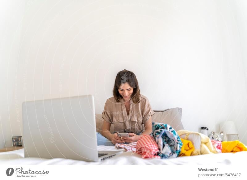 Lächelnde junge Frau benutzt Mobiltelefon auf dem Bett, umgeben von Kleidung Badebekleidung Bikinis Hemden Betten Rechner Laptops Notebook Notebooks
