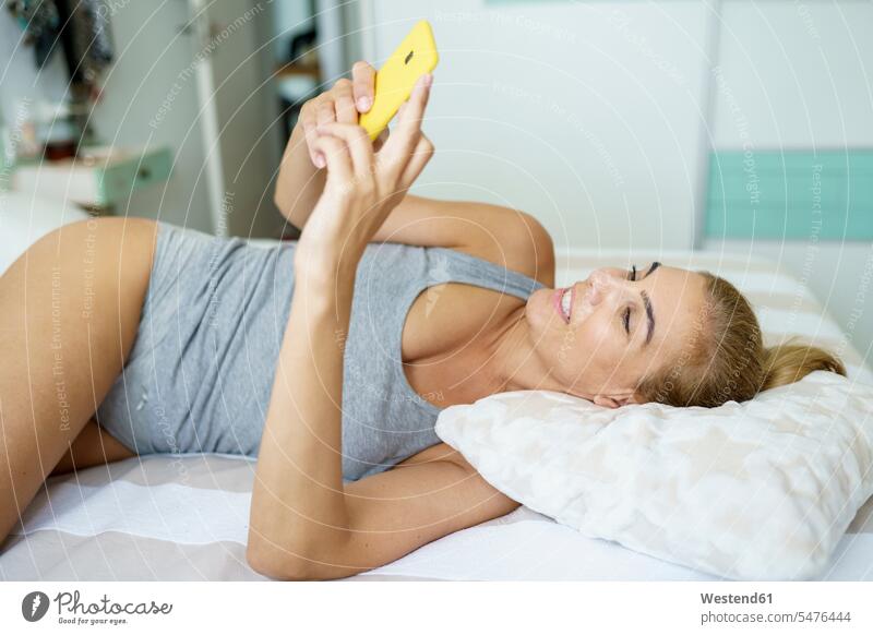 Lächelnde Frau sendet im Bett lächelnde Textnachrichten über Smartphone Farbaufnahme Farbe Farbfoto Farbphoto Spanien Innenaufnahme Innenaufnahmen innen drinnen