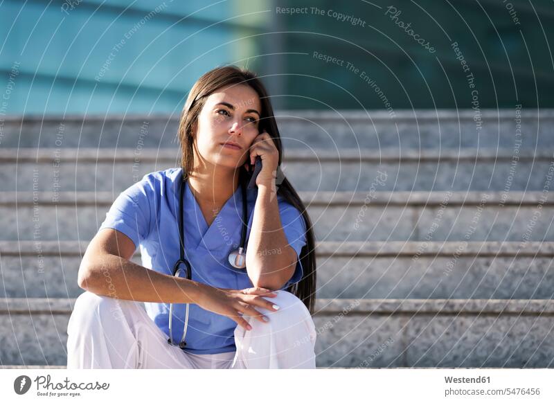 Junger Arzt telefoniert mit Handy, während er auf der Treppe vor dem Krankenhaus sitzt Farbaufnahme Farbe Farbfoto Farbphoto Außenaufnahme außen draußen