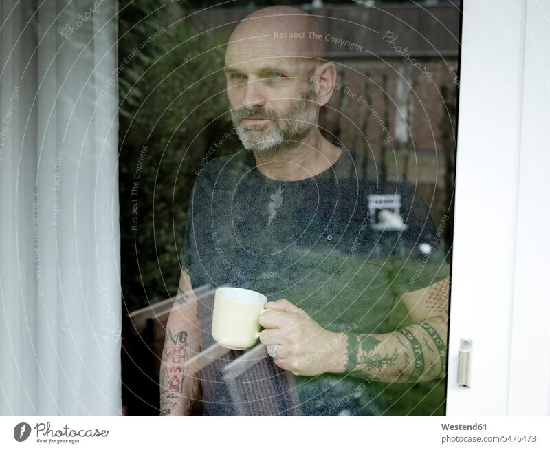 Tätowierter Mann, der Kaffee trinkt und aus dem Fenster schaut Zuhause zu Hause daheim aus dem Fenster sehen durchs Fenster schauen aus dem Fenster schauen
