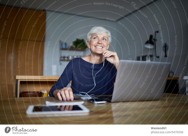 Aktive ältere Frau, die auf einem Smartphone Musik hört und einen Laptop benutzt, während sie von zu Hause weg schaut Farbaufnahme Farbe Farbfoto Farbphoto