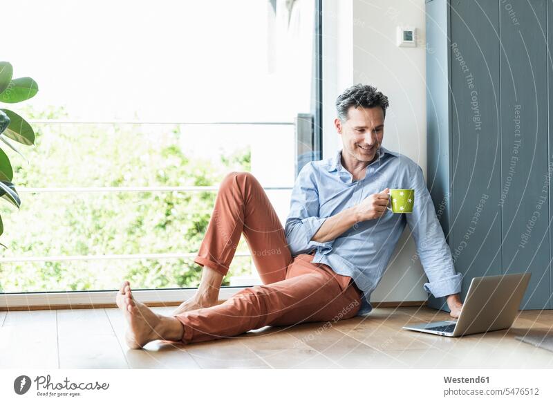 Reifer Mann entspannt sich zu Hause bei einer Tasse Kaffee, mit Laptop Rechner Laptops Notebook Notebooks entspannen relaxen sitzend sitzt freuen Frohsinn