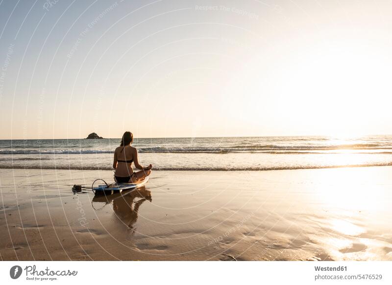 Junge Frau übt Yoga am Strand, sitzt auf Surfbrett, meditiert Meer Meere junge Frau junge Frauen Schneidersitz Yoga-Übungen Yogauebungen Yogaübungen Jogauebung