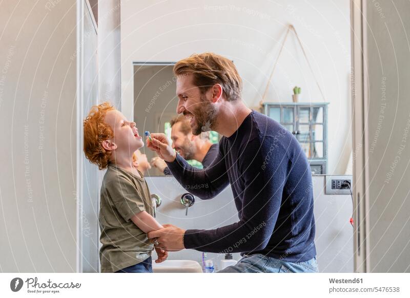 Glücklicher Mann putzt dem Sohn zu Hause im Badezimmer die Zähne mit der Zahnbürste Farbaufnahme Farbe Farbfoto Farbphoto Innenaufnahme Innenaufnahmen innen