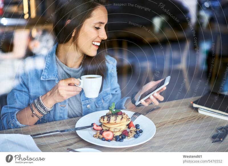 Lächelnde junge Frau mit Teller mit Pfannkuchen mit Telefon in Café lächeln Handy Mobiltelefon Handies Handys Mobiltelefone weiblich Frauen Pancake Eierkuchen