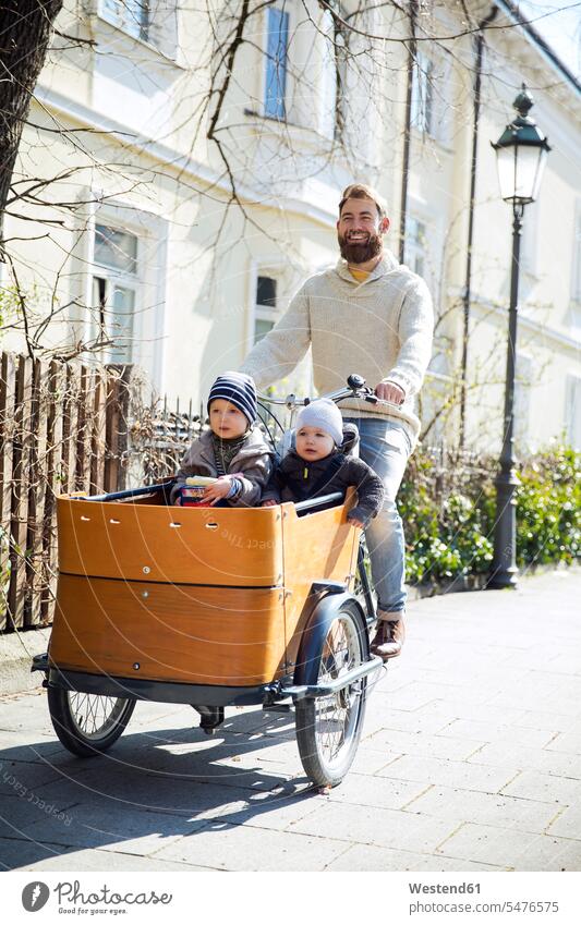 Glücklicher Vater mit zwei Kindern fahren Cargo Fahrrad in der Stadt Babies Babys glücklich glücklich sein glücklichsein Bikes Fahrräder Räder Rad Lastenfahrrad