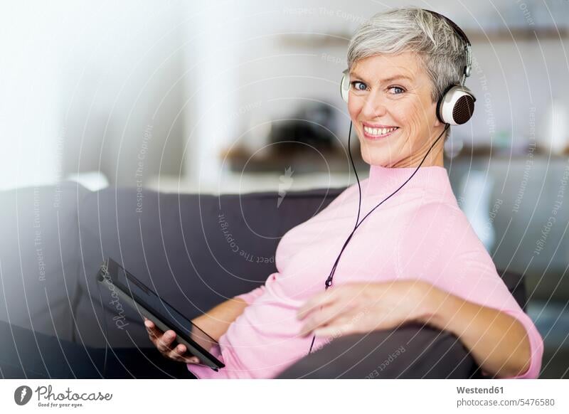 Porträt der glücklichen reifen Frau sitzt auf der Couch mit digitalen Tablette hören Musik mit Kopfhörern sitzen sitzend Portrait Porträts Portraits weiblich