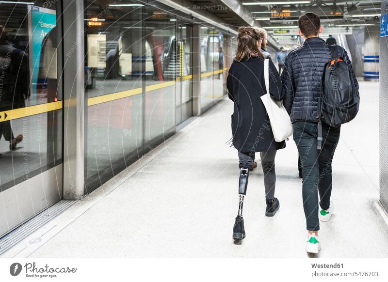 Rückansicht einer jungen Frau mit Beinprothese und eines gehenden Mannes auf dem Bahnsteig einer U-Bahn-Station Freunde Kameradschaft Rucksäcke Taschen Jacken