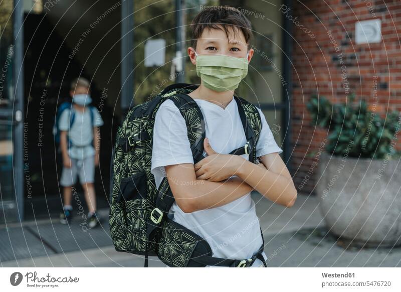 Kleiner Junge mit schützender Gesichtsmaske und gekreuzten Armen vor dem Schulgebäude stehend Farbaufnahme Farbe Farbfoto Farbphoto Außenaufnahme außen draußen