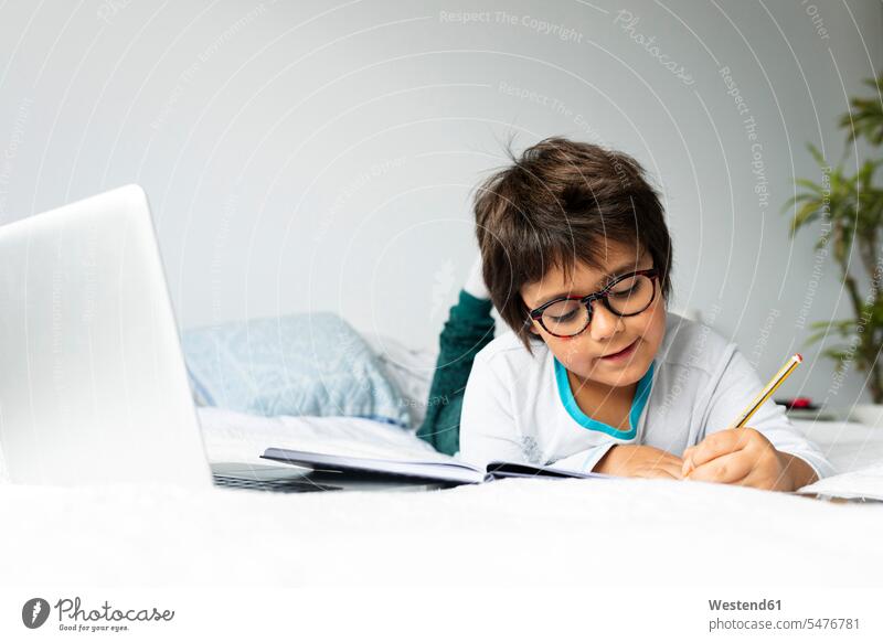 Porträt eines kleinen Jungen, der mit einem Laptop auf dem Bett liegt und Hausaufgaben macht Ausbildung Schueler Schulkinder Schüler Betten Stifte Bleistifte