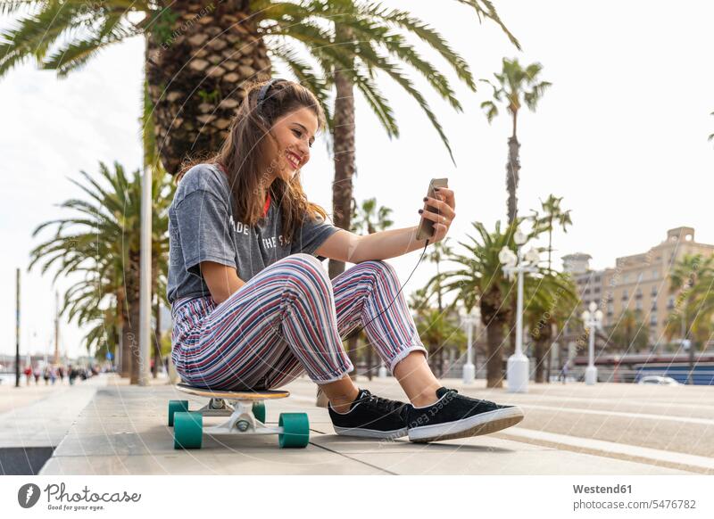 Glückliches Teenager-Mädchen sitzt auf einem Skateboard und hört Musik mit Kopfhörern Rollbretter Skateboards glücklich glücklich sein glücklichsein hören