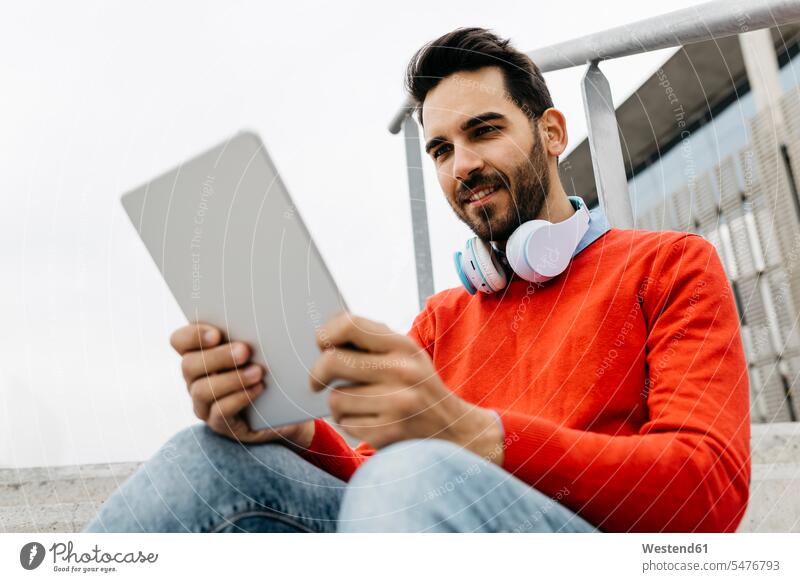 Lässiger Geschäftsmann auf einer Treppe in der Stadt, der sein digitales Tablet und seine Kopfhörer benutzt benutzen benützen Businessmann Businessmänner