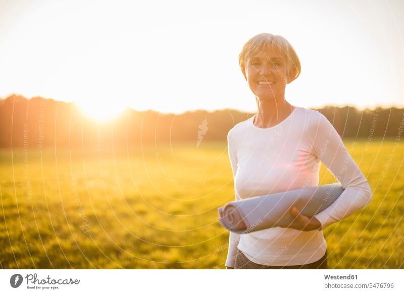 Porträt einer lächelnden älteren Frau, die eine Yogamatte auf einer ländlichen Wiese bei Sonnenuntergang hält auf dem Land auf dem Lande Portrait Porträts