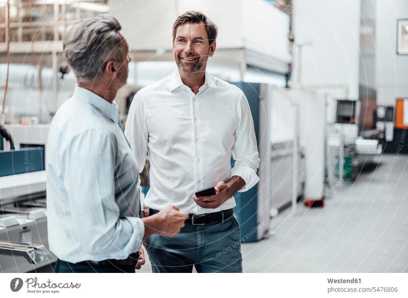 Ein reifer Geschäftsmann hält ein Smartphone in der Hand, während er mit einem Industriekollegen diskutiert Farbaufnahme Farbe Farbfoto Farbphoto Deutschland