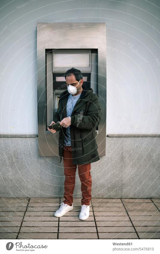 Mann mit Schutzmaske steht vor einem Geldautomaten und steckt Geld in seine Geldbörse Börse Börsen Geldboerse Geldboersen Geldbörsen Portemonnaies Portmonee