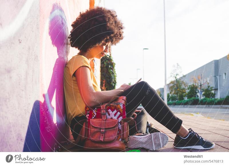 Junge Frau mit Afrofrisur sitzt an Graffiti-Wand und benutzt Smartphone Leute Menschen People Person Personen gelockt gelockte Haare gelocktes Haar lockig