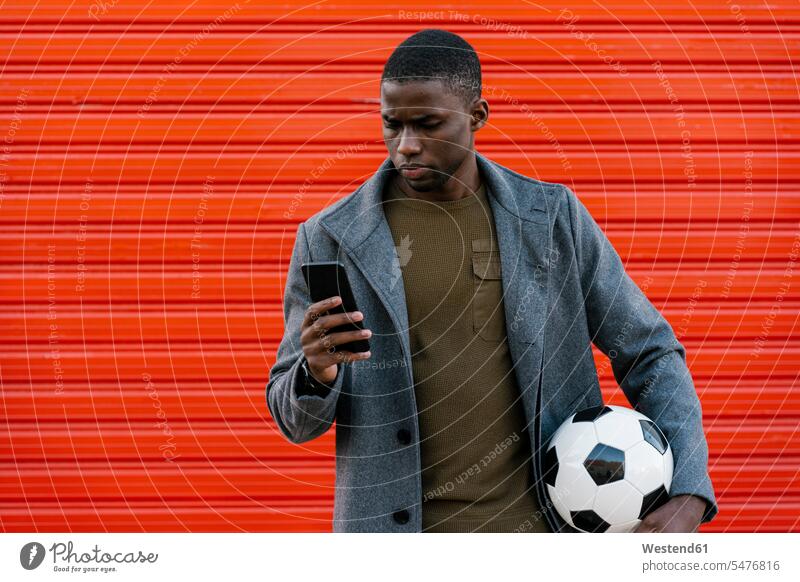 Junger Mann mit Fußball, der ein Mobiltelefon benutzt, während er an einer roten Wand steht Farbaufnahme Farbe Farbfoto Farbphoto Außenaufnahme außen draußen
