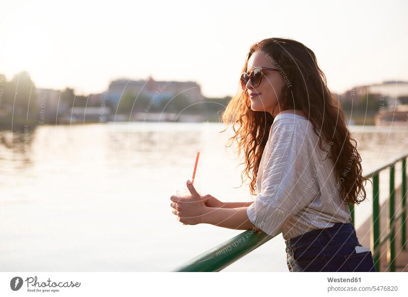 Lächelnde junge Frau mit einem Getränk zum Mitnehmen, die den Sonnenuntergang am Flussufer genießt Getraenk Getränke Getraenke Sonnenuntergänge genießen