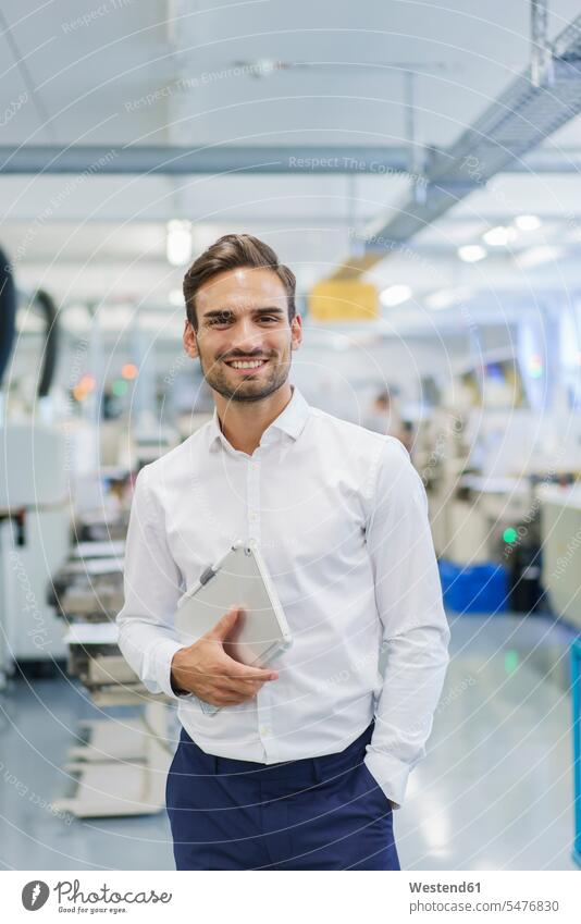 Lächelnder junger Geschäftsmann hält digitales Tablett in der Hand, während er mit den Händen in den Taschen in der beleuchteten Fabrik steht Farbaufnahme Farbe