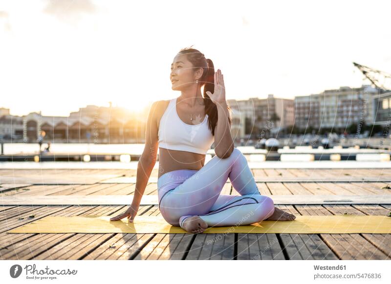Asiatische Frau praktiziert Yoga auf einem Pier im Hafen, halbe Wirbelsäulenverdrehung bei Sonnenuntergang sitzend sitzt abends farbig mehrfarbig weiss weiße