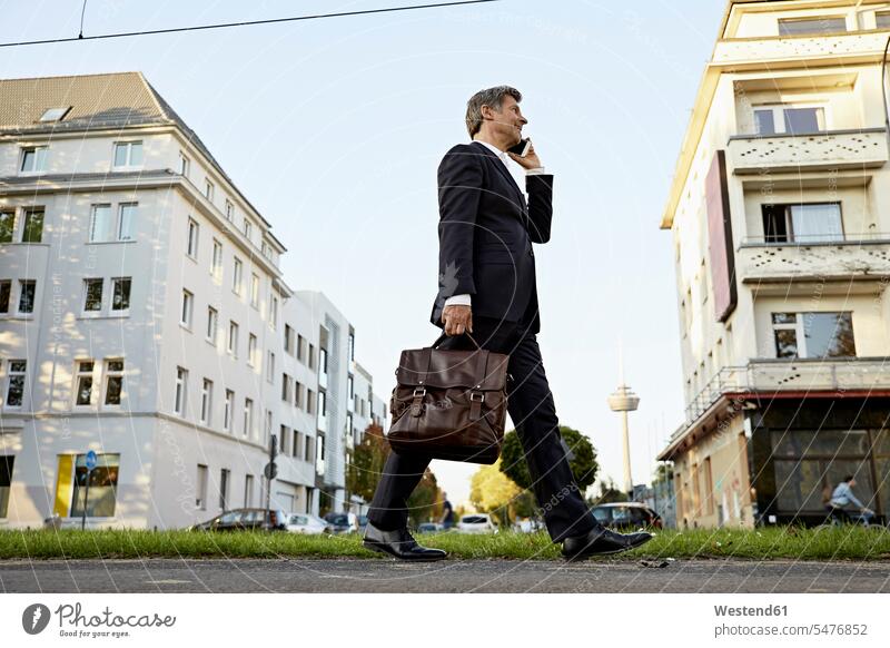 Reifer Geschäftsmann zu Fuß und mit Smartphone in der Stadt Businessmann Businessmänner Geschäftsmänner gehen gehend geht staedtisch städtisch iPhone