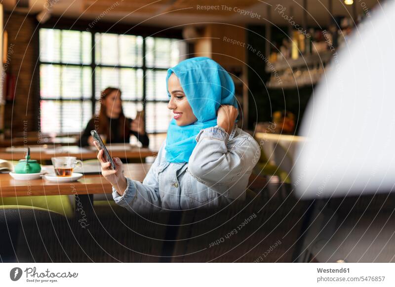 Junge Frau trägt einen türkisfarbenen Hijab und benutzt ein Smartphone in einem Cafe Leute Menschen People Person Personen Europäisch Kaukasier kaukasisch