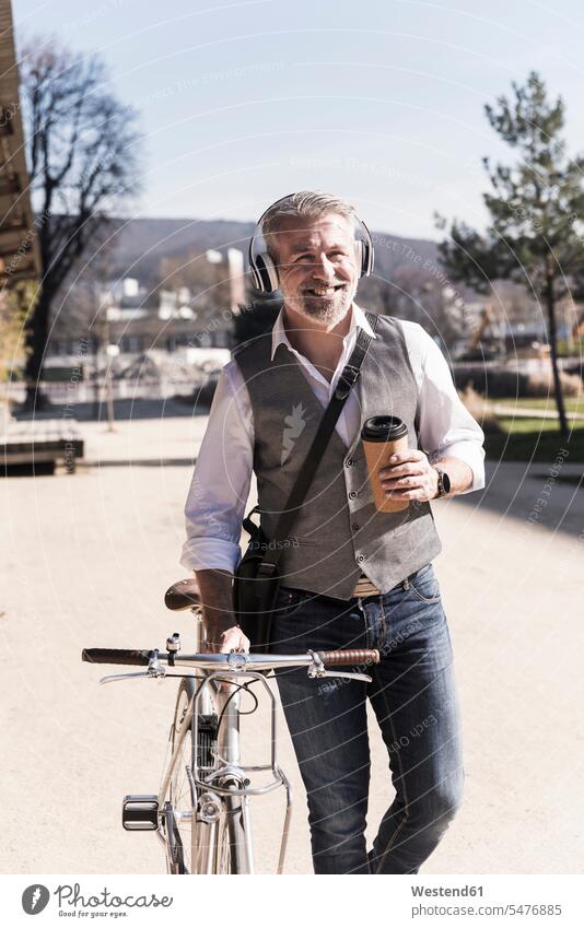 Lächelnder reifer Geschäftsmann mit Fahrrad, Kaffee zum Mitnehmen und Kopfhörern unterwegs in der Stadt lächeln Businessmann Businessmänner Geschäftsmänner