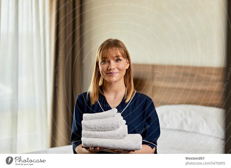 Lächelndes Zimmermädchen hält Handtücher, während es im Hotelzimmer steht Farbaufnahme Farbe Farbfoto Farbphoto Innenaufnahme Innenaufnahmen innen drinnen