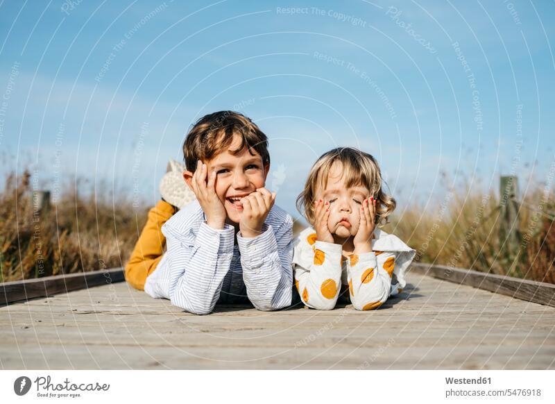 Porträt eines Jungen und seiner kleinen Schwester, die nebeneinander auf der Promenade liegen und lustige Gesichter ziehen Harmonie harmonisch Seite an Seite