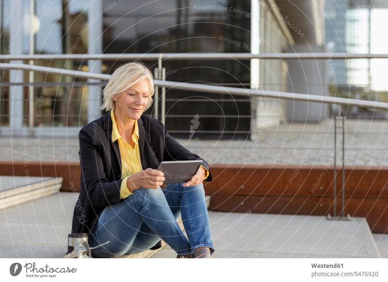 Lächelnde ältere Geschäftsfrau sitzt mit Tablette in der Stadt Zuversicht Zuversichtlich Selbstvertrauen selbstbewusst Vertrauen sitzen sitzend lächeln