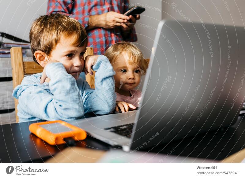 Bruder und Schwester schauen ein Video auf einem Laptop, Vater mit Smartphone im Hintergrund Junge Buben Knabe Jungen Knaben männlich Mädchen weibliche Babys