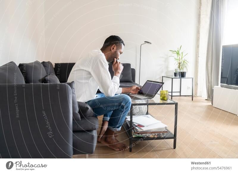 Mann sitzt auf Couch in moderner Wohnung und benutzt Laptop Bücher Couches Liege Sofas Rechner Laptops Notebook Notebooks sitzend daheim zu Hause Muße Technik