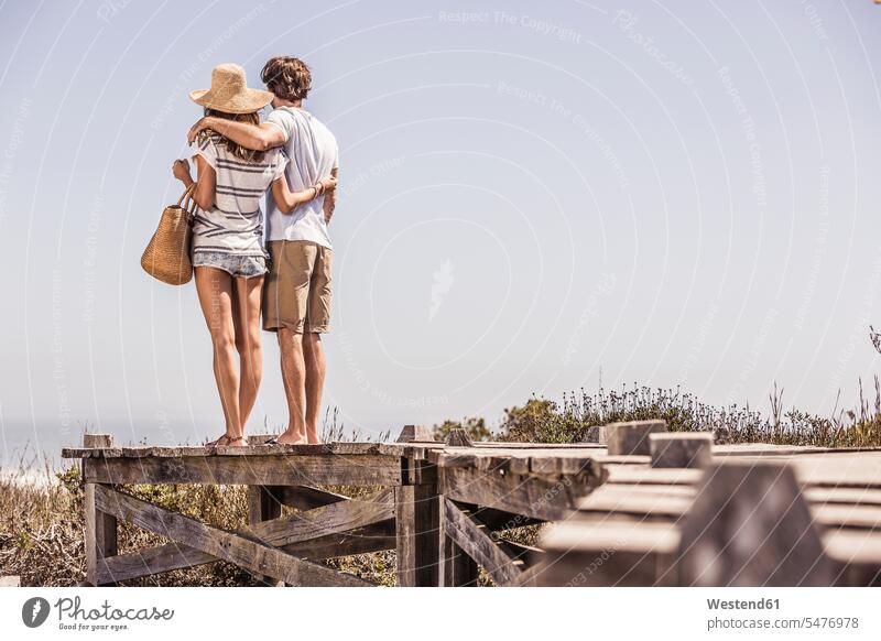 Junges Paar, das auf einer Strandpromenade an der Küste steht und die Aussicht betrachtet Touristen Taschen Arm umlegen Umarmung Umarmungen Jahreszeiten