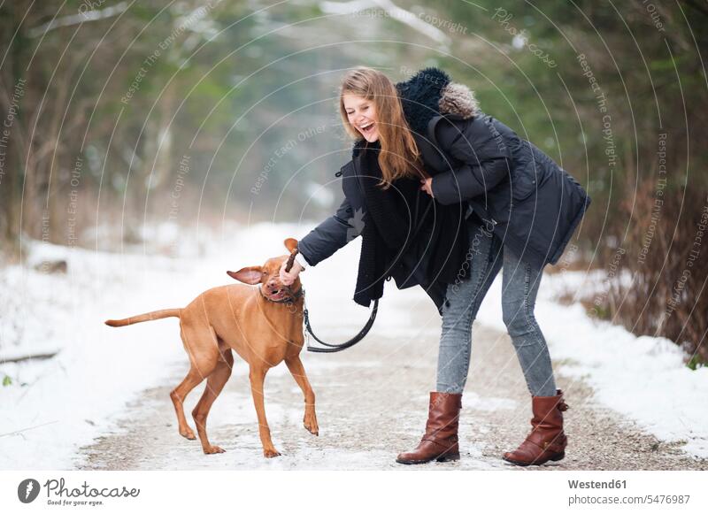 Fröhliche junge Frau spielt im Winter mit Hund auf Waldstrasse Farbaufnahme Farbe Farbfoto Farbphoto Freizeitbeschäftigung Muße Zeit Zeit haben Außenaufnahme