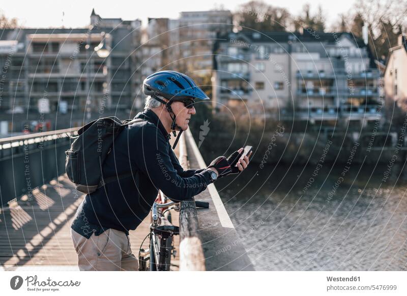 Älterer Mann mit Radfahrerhelm mit Smartphone auf der Brücke radfahren fahrradfahren radeln Senior ältere Männer älterer Mann Senioren iPhone Smartphones