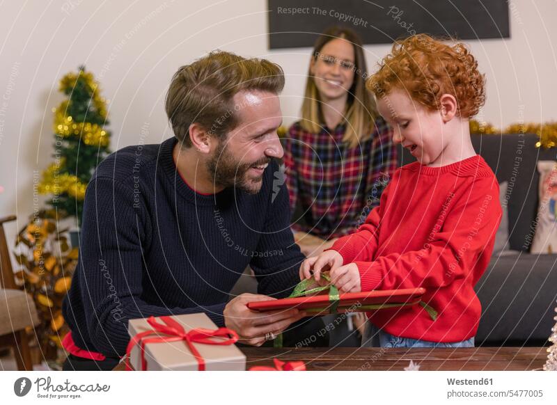 Lächelnde Frau schaut den Vater an, der dem Sohn im Wohnzimmer ein Weihnachtsgeschenk macht Farbaufnahme Farbe Farbfoto Farbphoto Freizeitbeschäftigung Muße