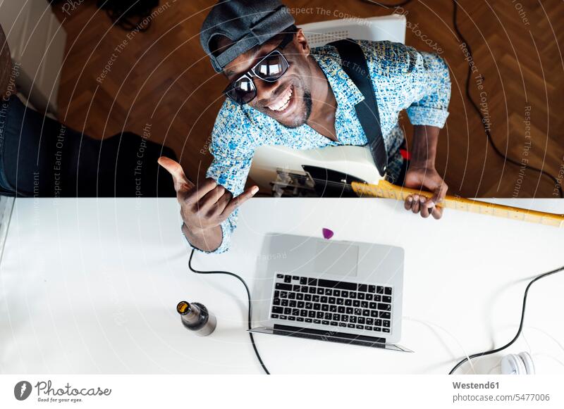 Porträt eines lächelnden jungen Mannes mit Shaka-Zeichen, der mit Gitarre und Laptop am Schreibtisch sitzt Farbaufnahme Farbe Farbfoto Farbphoto Innenaufnahme