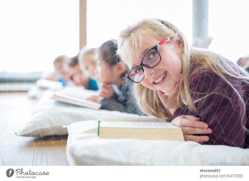 Porträt eines lächelnden Schulmädchens, das mit Klassenkameraden auf dem Boden liegt und im Pausenraum der Schule ein Buch liest lesen Lektüre Bücher liegen