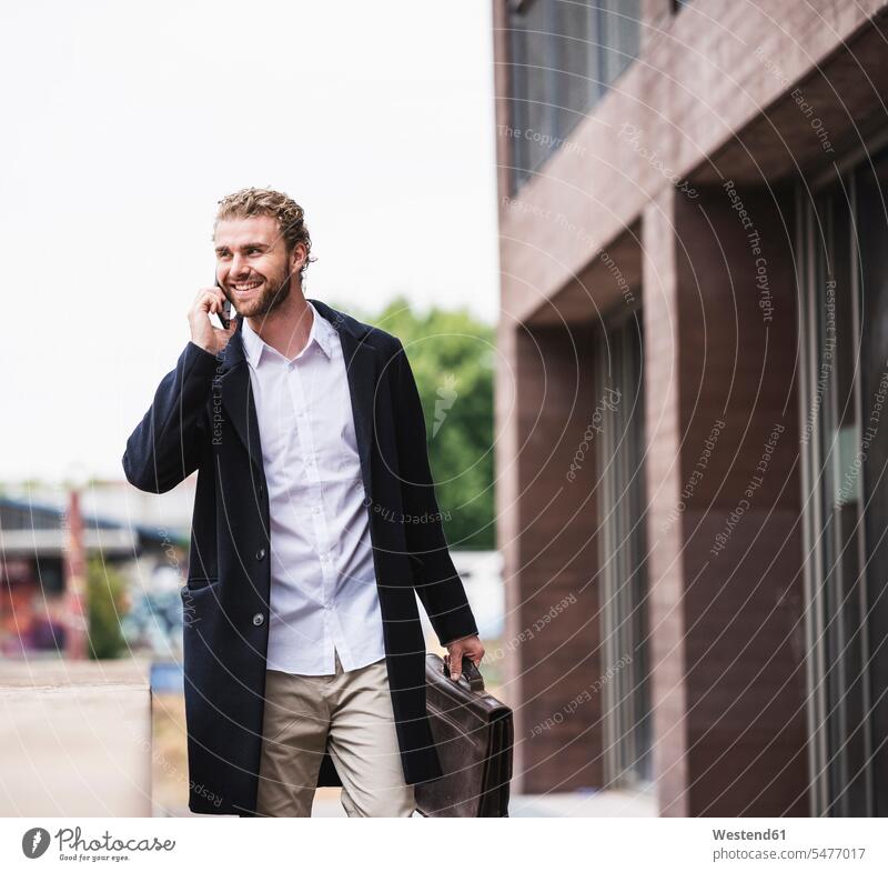 Lächelnder junger Geschäftsmann am Handy vor Bürogebäude Bürohaus Bürohäuser telefonieren anrufen Anruf telephonieren Mobiltelefon Handies Handys Mobiltelefone